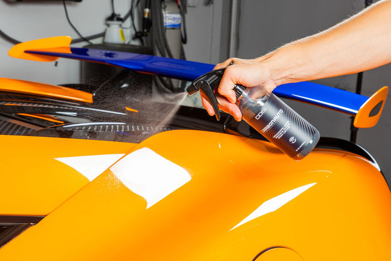Spraying a black bottle of CDS CarbonAuto Graphene Spray Coating protective paint over the orange body of a sports car in a garage setting.